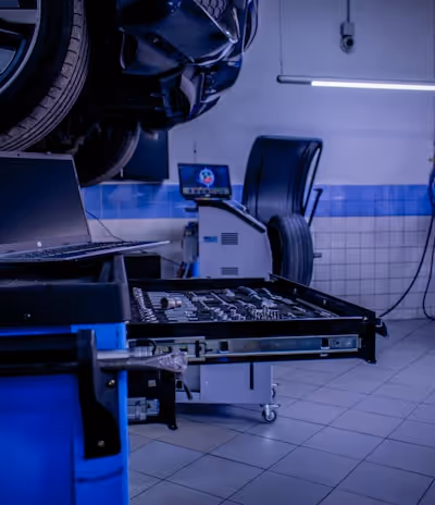 A car service or repair garage is depicted, with a vehicle hoisted on a lift. A laptop is placed on a nearby workstation with an open drawer filled with tools. The environment features tiled walls and floor, and equipment such as a tire changer and a balancing machine. Fluorescent lighting provides illumination.