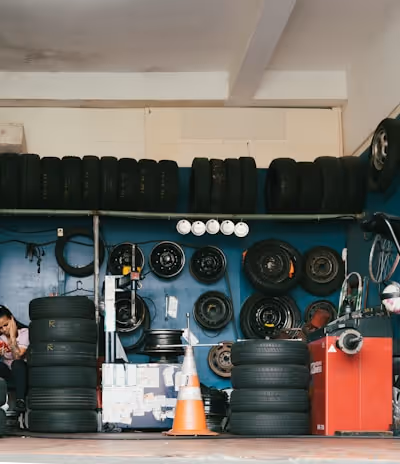 A garage filled with tires and automotive equipment. Tires are stacked on shelves and some are mounted on the walls alongside wheel rims. A person is seated in the corner on the left, looking at a mobile phone. An orange traffic cone is on the floor, and there is a machine used for tire services.
